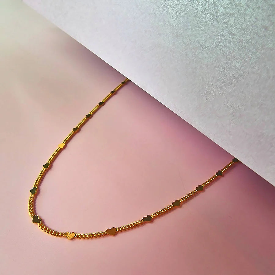 A heart-shaped necklace with gold chain links rests on a pink surface, partly covered by a white sheet—ideal as a stylish and hypoallergenic accessory for everyday wear.
