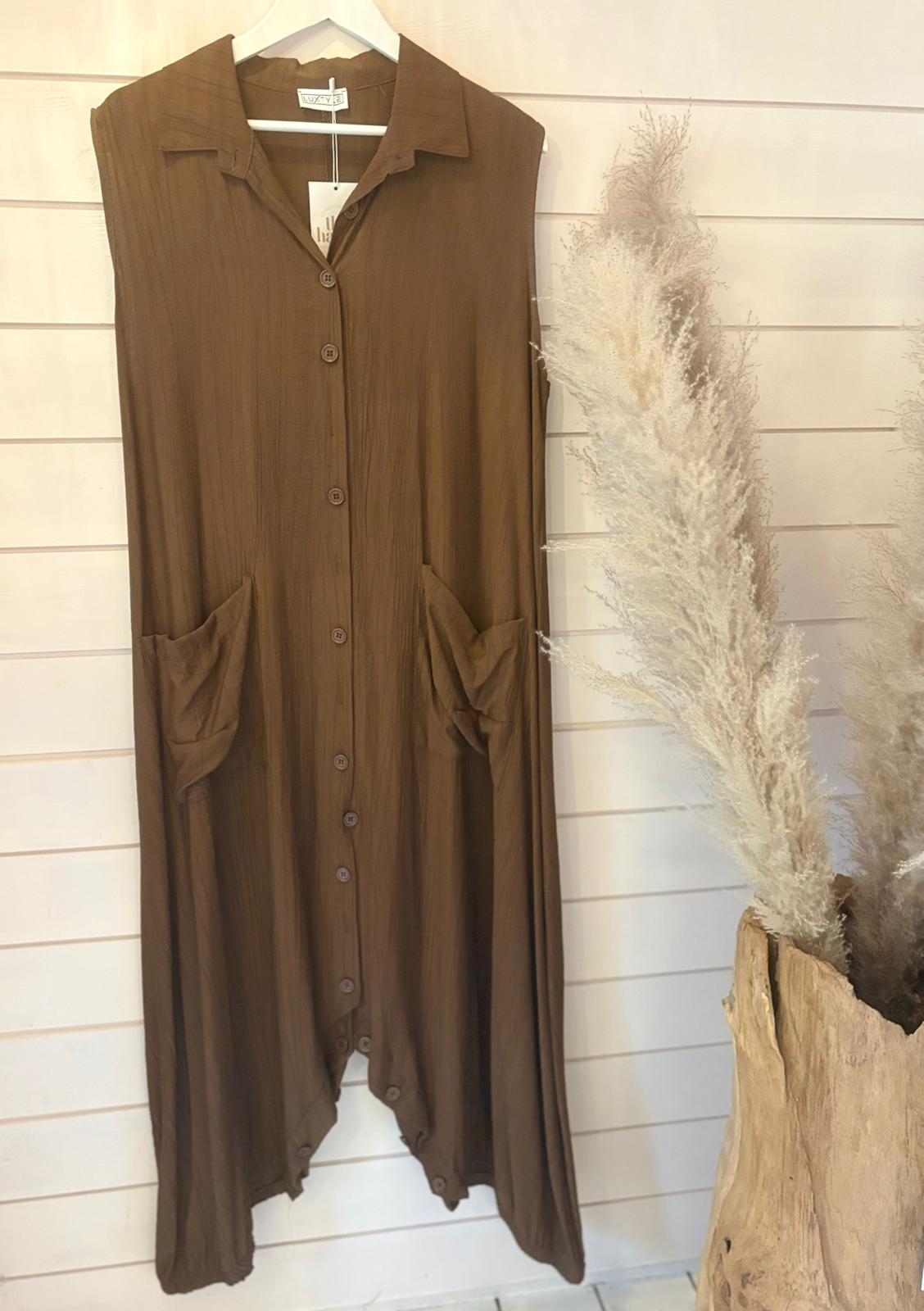 Sleeveless brown jumpsuit dress with a long silhouette and large front pockets hangs on a white wall, beside a vase containing dried pampas grass.