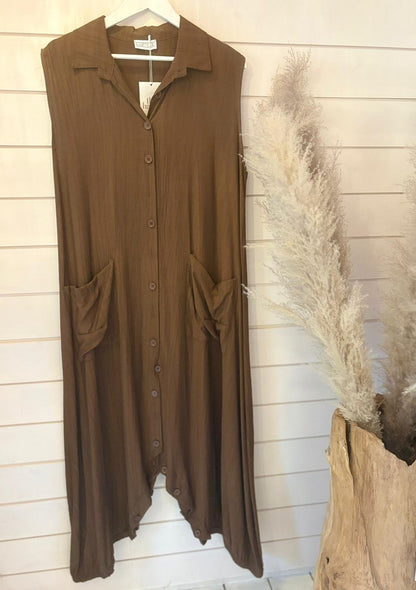 Sleeveless brown jumpsuit dress with a long silhouette and large front pockets hangs on a white wall, beside a vase containing dried pampas grass.