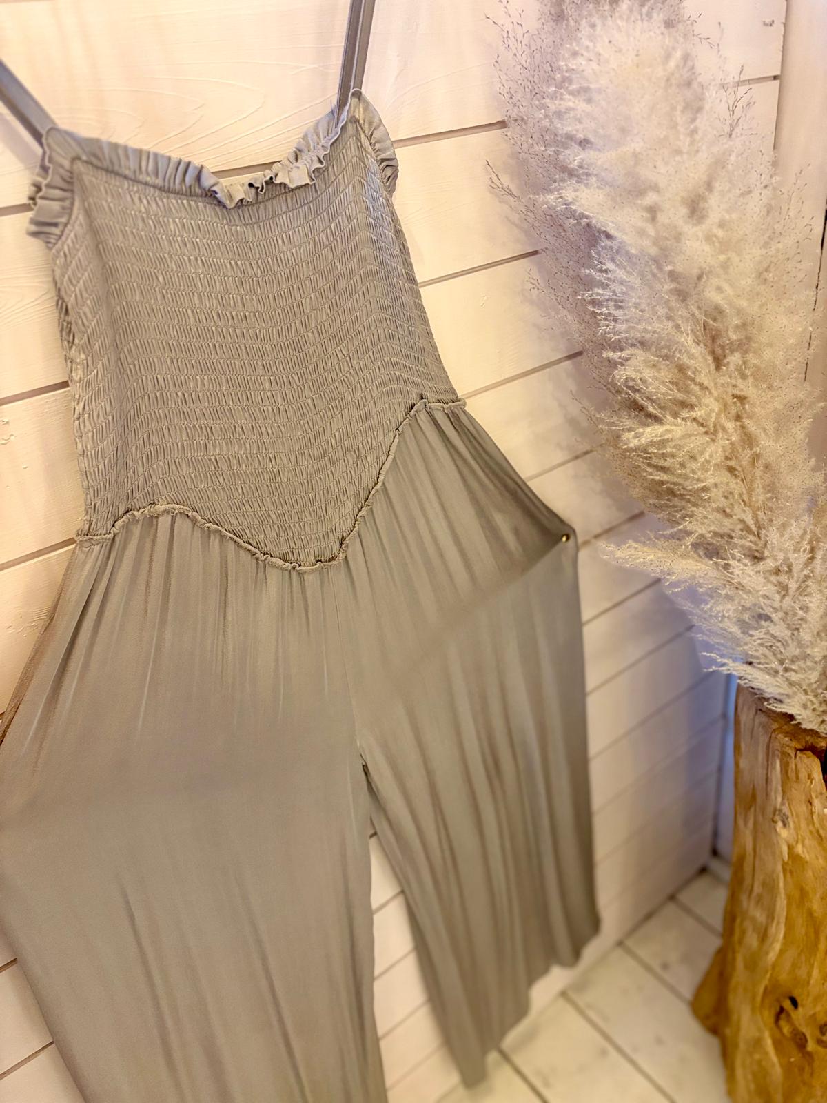 Light grey sleeveless jumpsuit with a smocked bodice and wide legs hanging against a white wooden wall, beside a vase of pampas grass.