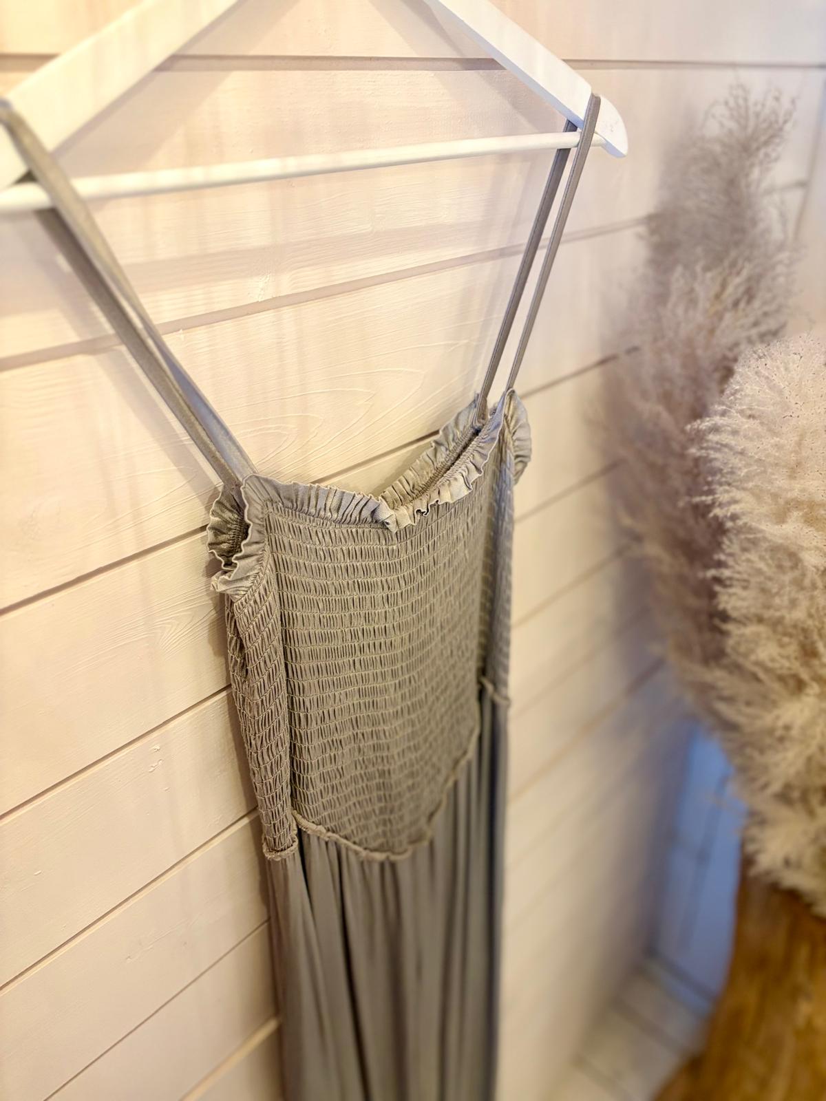A light grey, sleeveless, smocked dress with thin straps hangs on a white hanger against a wooden wall, with pampas grass décor nearby.
