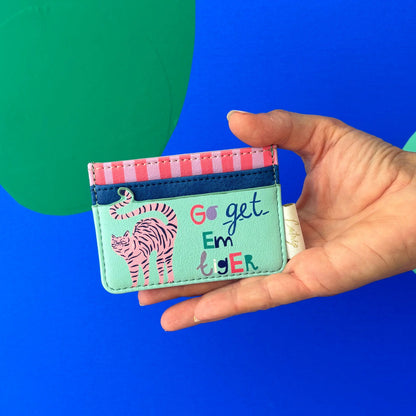 A hand holds a cat cardholder adorned with cheerful cat illustrations, including a tiger and the phrase "Go get em tiger," set against a blue and green background.