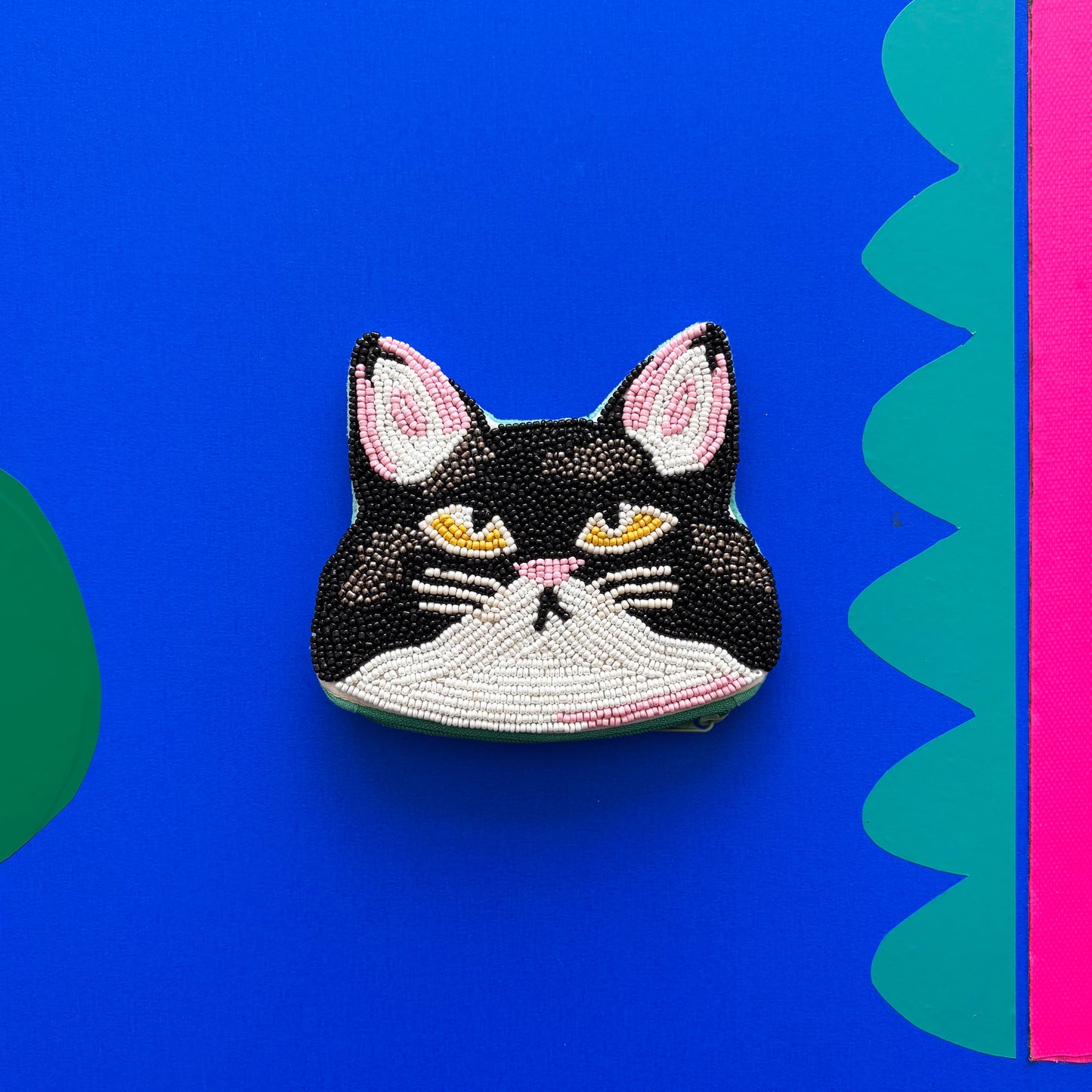 A beaded black and white cat face accessory pin with yellow eyes, set against a bright blue background featuring green and pink shapes—a perfect gift for cat lovers.