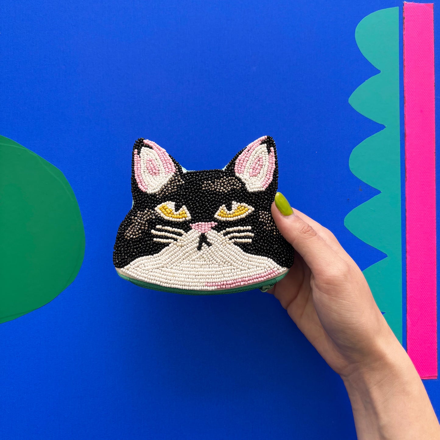 A hand holds a beaded cat coin purse shaped like a black and white kitty face, set against a bright blue background with green and pink geometric shapes.