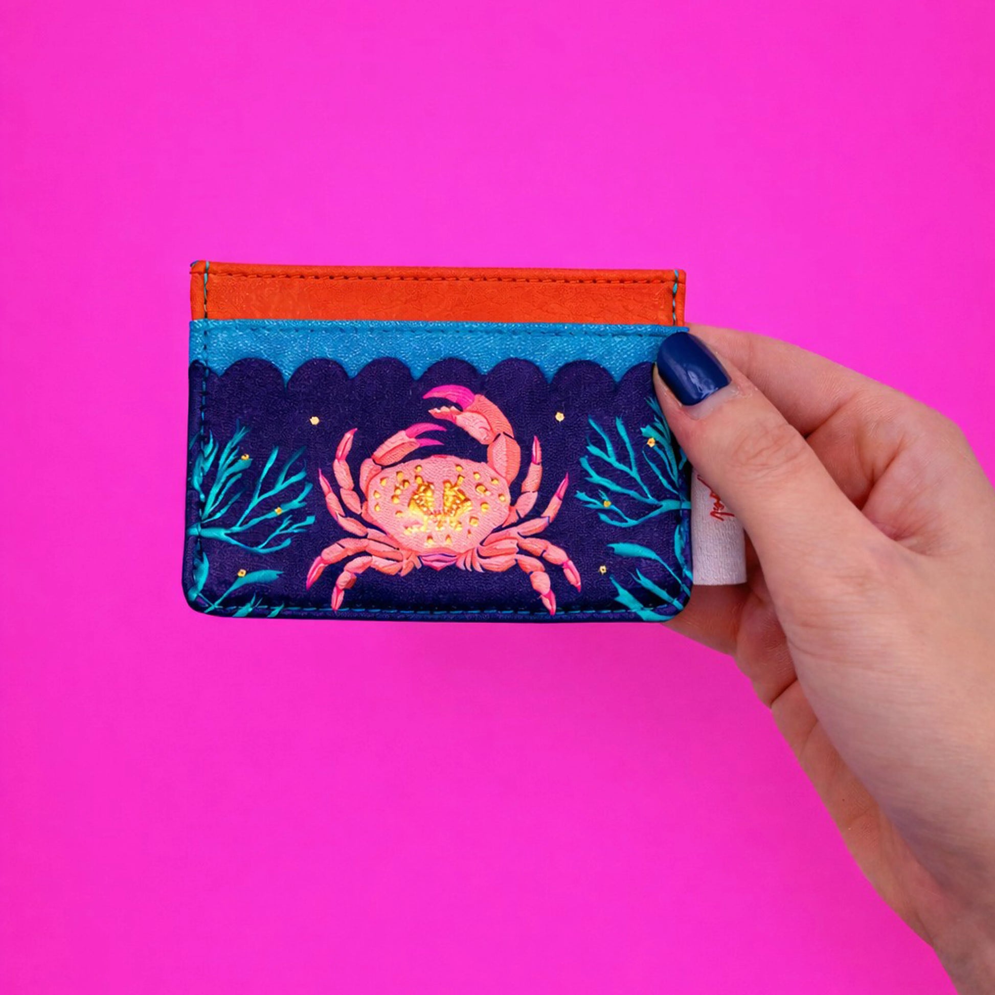 A hand holding a colourful card holder featuring a crab and seaweed design, set against a vivid pink background.