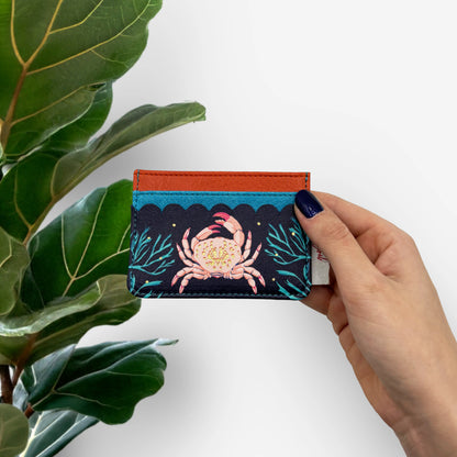 A hand holding a colourful Crab card holder wallet featuring a crab illustration, positioned beside a green leafy plant on a white background.