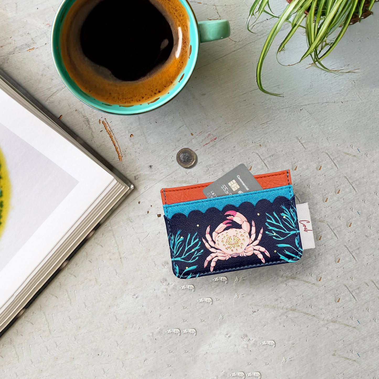 A turquoise cup of coffee, an open book, a coin, and a colourful card holder with cards rest on a grey tabletop beside a small plant.