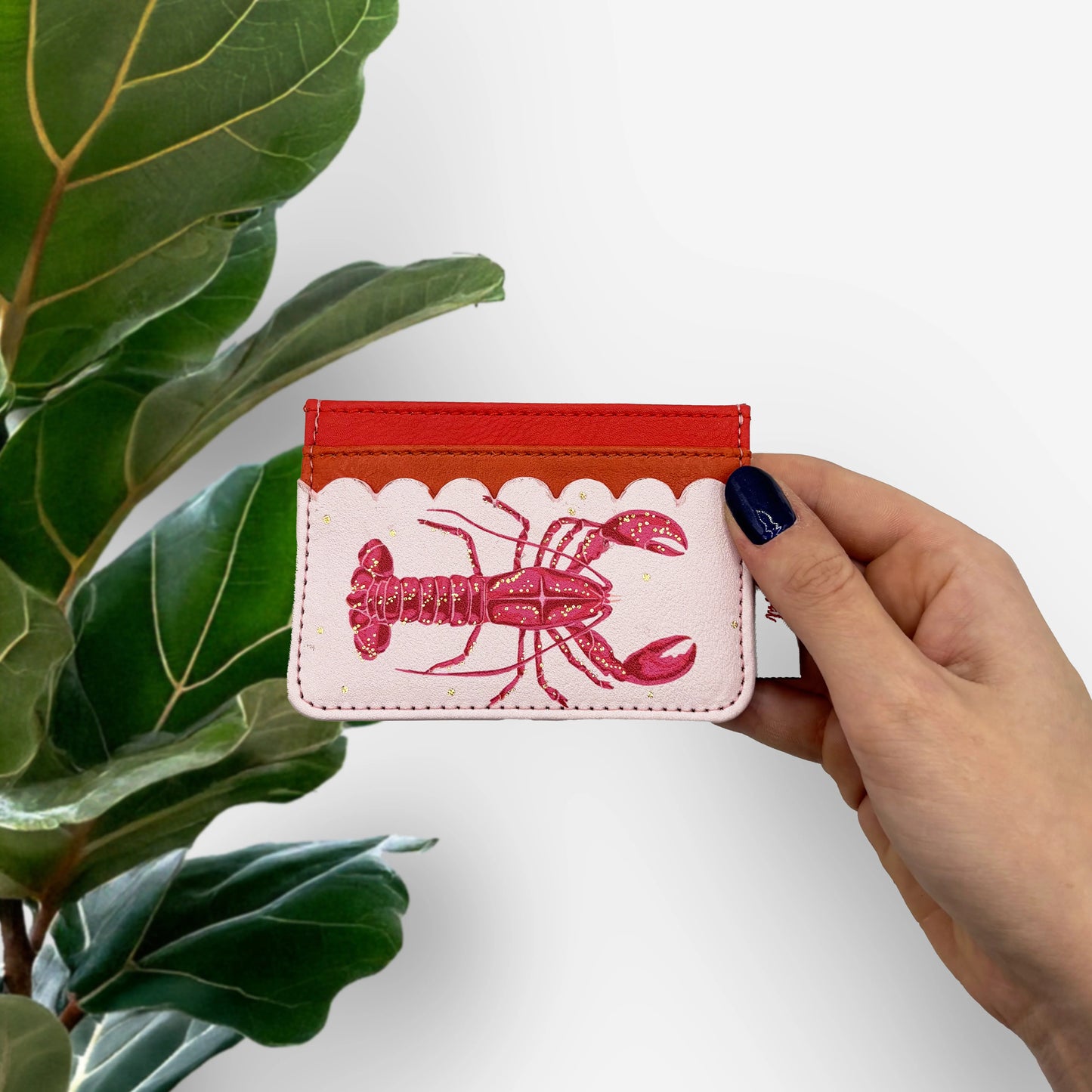 A hand with dark blue nail varnish holds a Coral Lobster Card Holder crafted from durable PVC, displaying a red lobster illustration and card slots, positioned beside a green leafy plant.