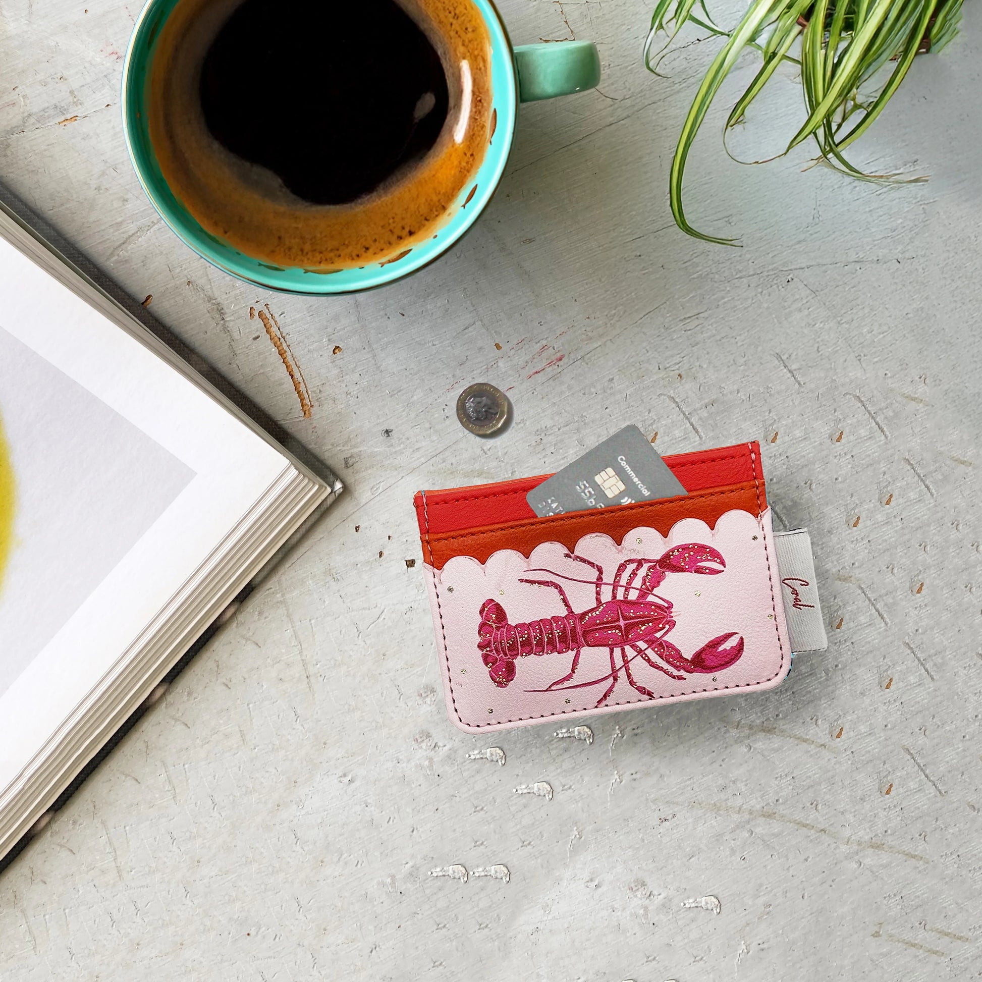 A coffee cup, a book, a plant and a coin rest on a table beside a Coral Lobster Card Holder crafted from durable PVC, featuring handy card slots and a vibrant red lobster design.