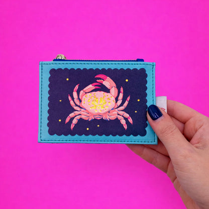 A hand with dark blue nail varnish holds a Coral Crab Zip Purse featuring a cheerful crab design, set against a vibrant pink background.