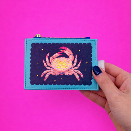 A hand with dark blue nail varnish holds a Coral Crab Zip Purse featuring a cheerful crab design, set against a vibrant pink background.