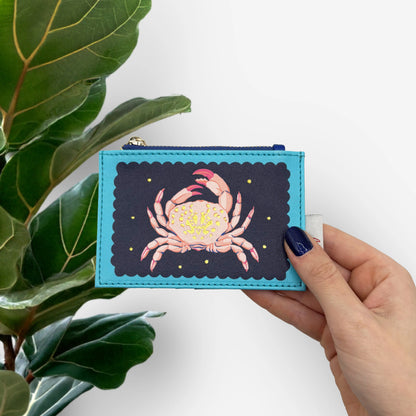 A hand holds a blue wallet with a delightful crab design beneath yellow stars, set against a white background with green leaves visible on the left.