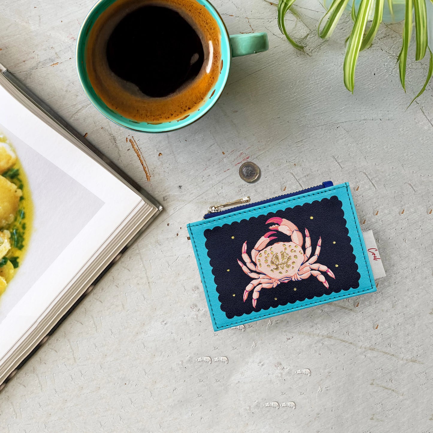 A teal mug of coffee, an open book, a plant, and a Coral Crab Zip Purse adorned with a delightful crab motif are arranged on a light grey, textured surface.