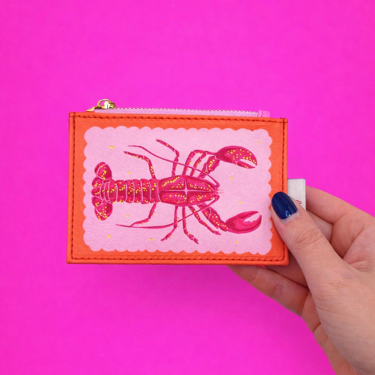 A hand holds a compact zipped purse adorned with a striking lobster motif against a vibrant pink backdrop—the Coral Lobster Zip Purse adds playful charm to your accessories.