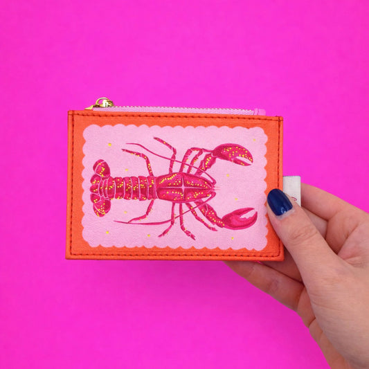A hand holds a compact zipped purse adorned with a striking lobster motif against a vibrant pink backdrop—the Coral Lobster Zip Purse adds playful charm to your accessories.