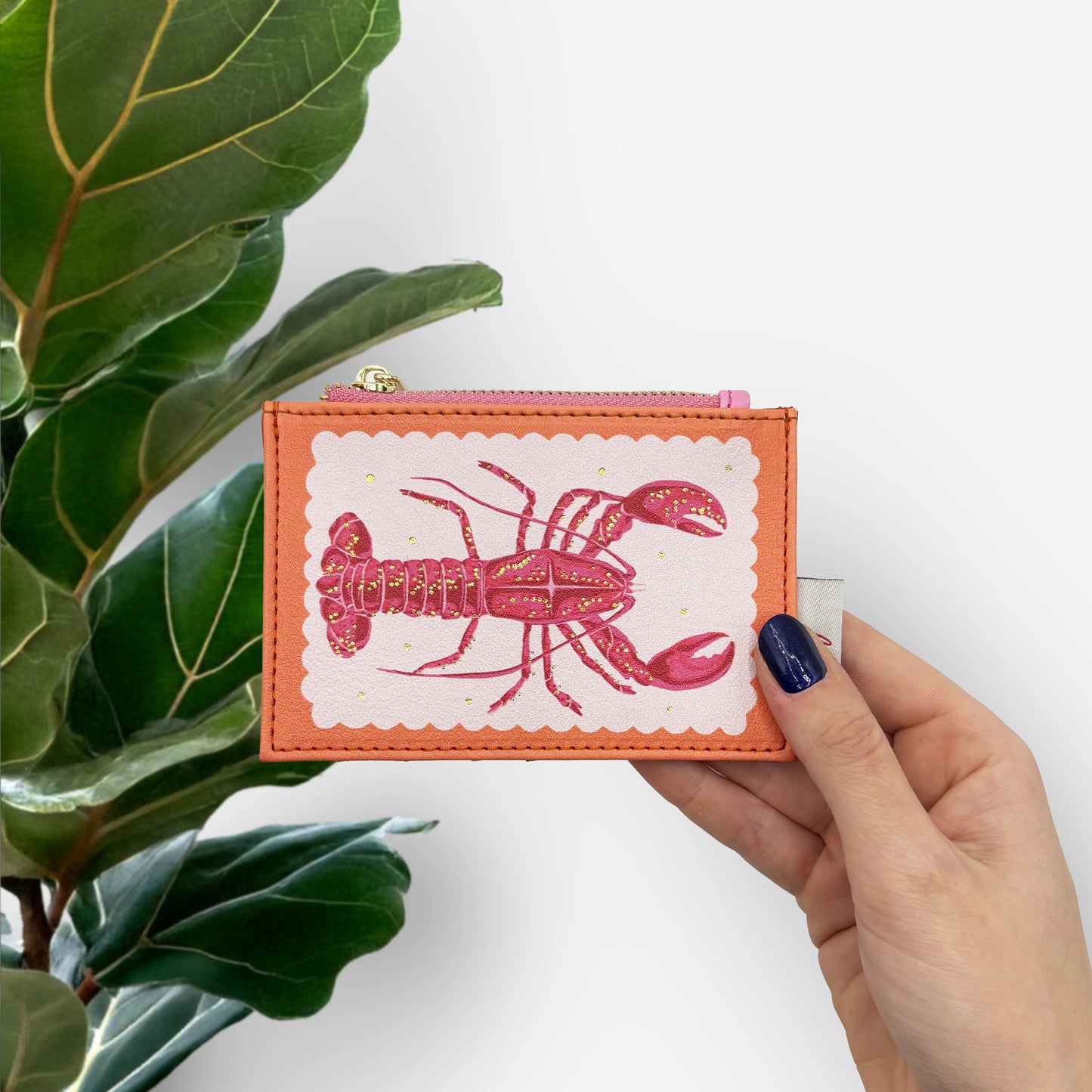 A hand holds a Coral Lobster Zip Purse featuring a pink lobster illustration, beside a green leafy plant set against a white background.