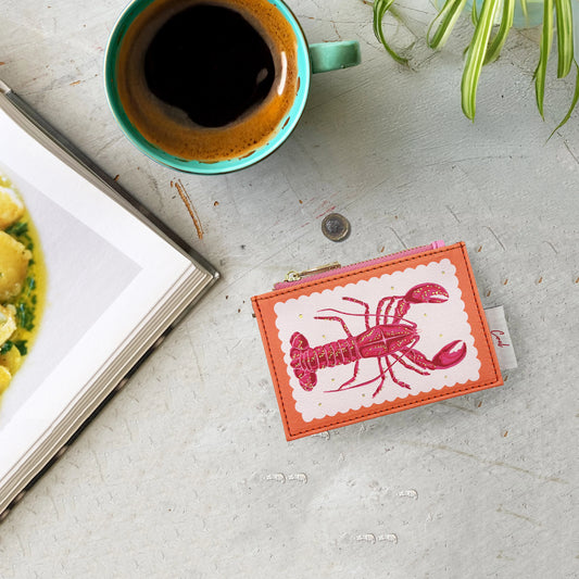 A teal mug of black coffee, an open book, a Coral Lobster Zip Purse, a coin, and a plant are placed on a light grey surface.