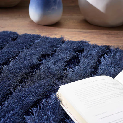 An open book sits on a soft, navy blue shag pile rug, with blurred ceramic vases and wooden flooring visible in the background.