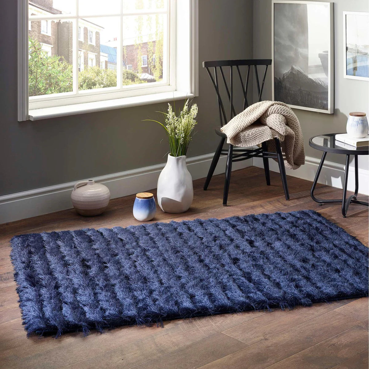 A blue textured, cruelty-free rug lies on a wooden floor in a bright room featuring a window, black chair, vases, blanket, and framed artwork.