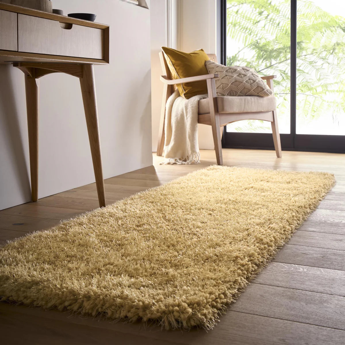 A beige hand-tufted shaggy rug on a wooden floor beside a wooden chair with cushions and a blanket, near a sliding glass door letting in natural light.