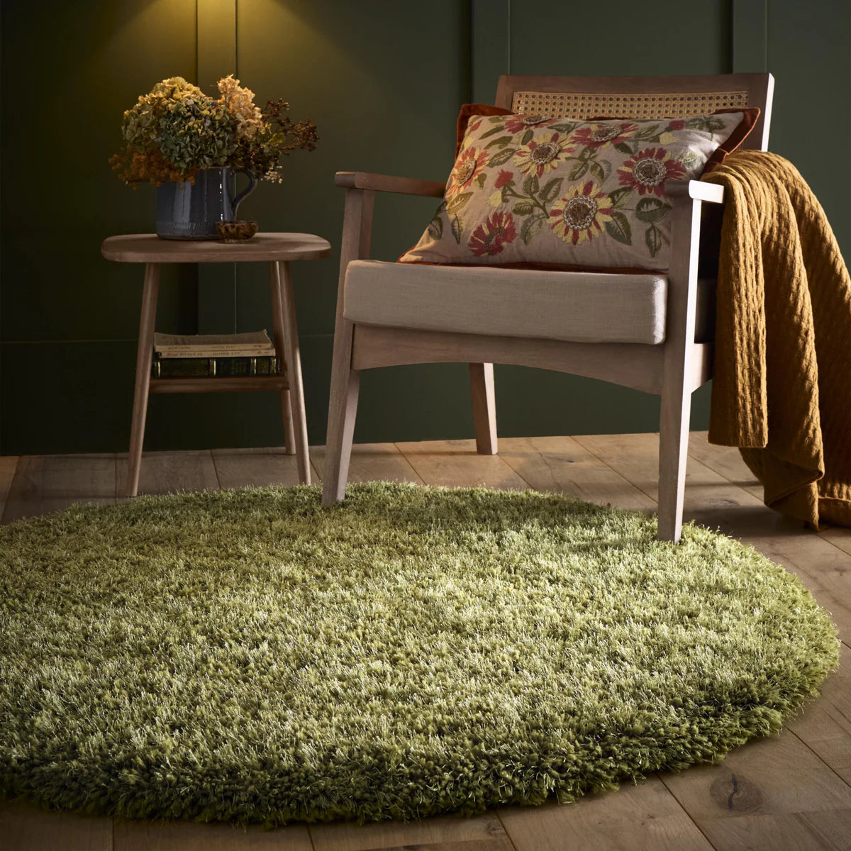 A wooden chair with a floral cushion and yellow throw sits next to a small round table with a vase of flowers on a hand-tufted Chicago rug with green shaggy pile, in a warmly lit room with green walls.