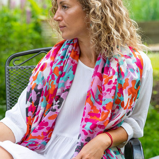A woman with curly blonde hair sits on a metal chair outdoors, wearing a white dress and a lightweight scarf made from vegan materials.