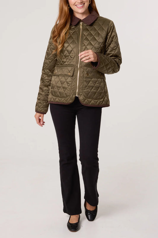A woman in a vegan khaki diamond-quilted coat, black trousers, and black shoes stands against a plain background.
