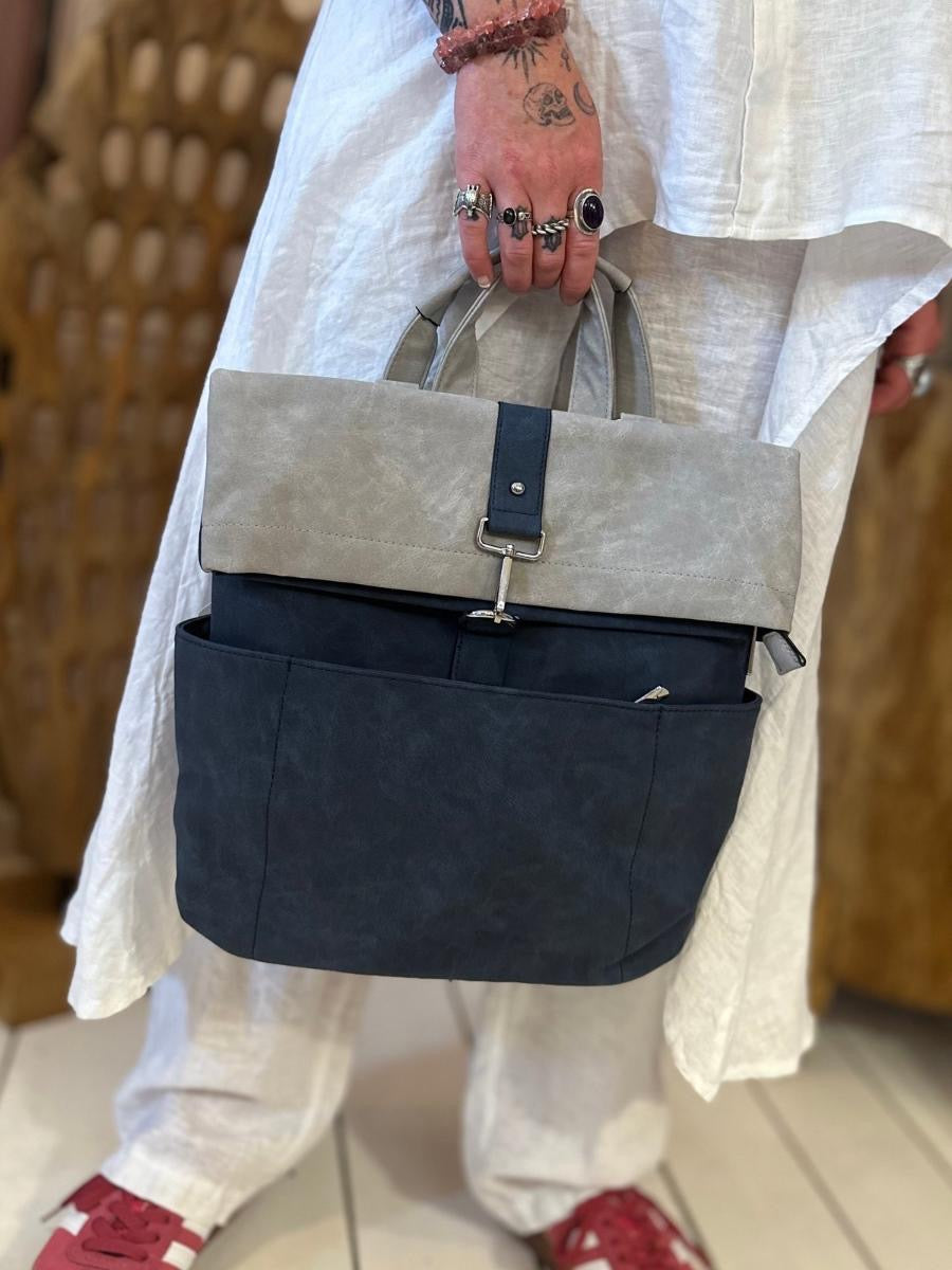 Person in a white outfit holding a two-tone grey rucksack with a buckle and outer pockets; only their hand, the bag, and part of their red trainers are visible.