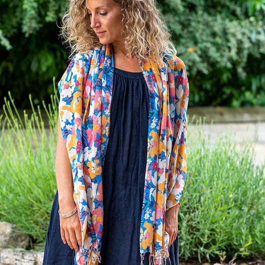 A woman with curly hair wears a dark dress and a lightweight summer scarf featuring a vibrant floral pattern while standing outdoors in front of greenery.