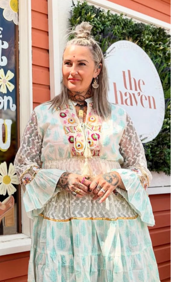 A woman with tattoos and grey hair styled half-up stands outside a building, wearing a floral-patterned Jessie Short Dress featuring mesh details and elegant bell sleeves.