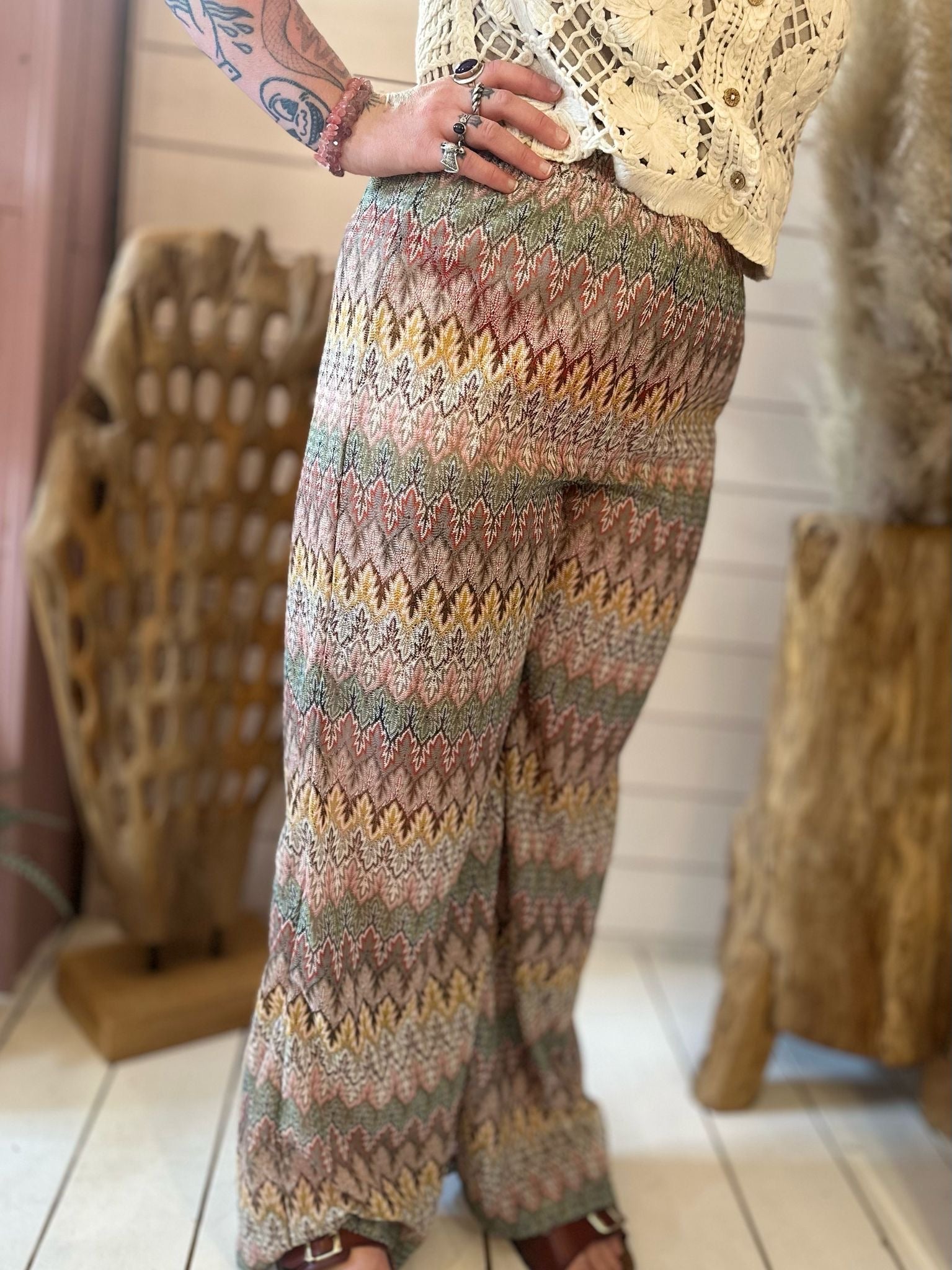 Person wearing Aztec mesh trousers with multi-coloured patterns, a cream crochet top, and sandals, standing indoors on a white floor next to wooden décor.