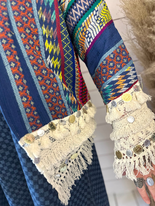 A close-up of a person wearing a blue patterned garment with multicoloured designs and cream-coloured lace cuffs adorned with coins and tassels.