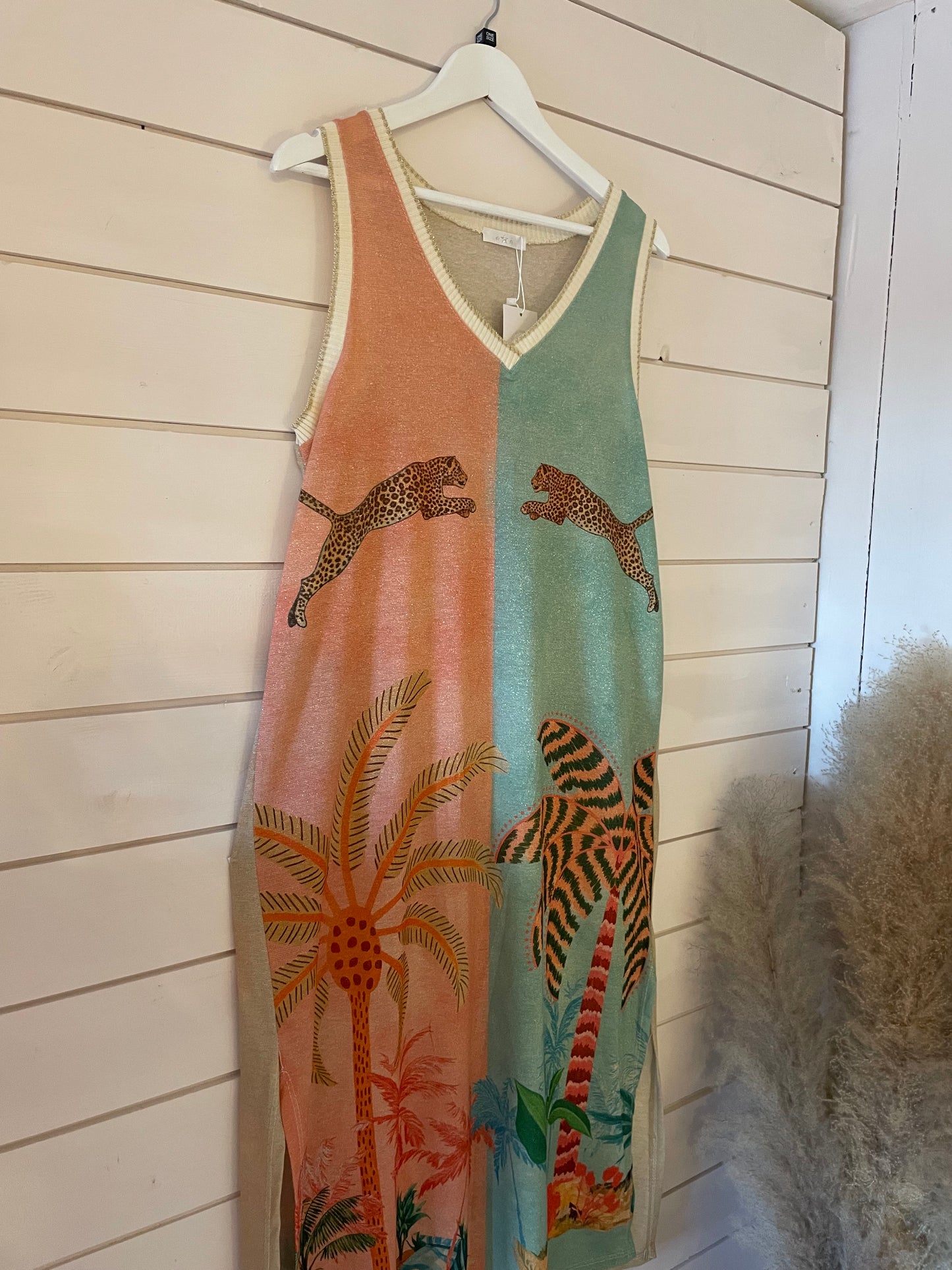 Sleeveless maxi dress with a tropical print, showcasing palm trees and leaping leopards, made from vegan materials and displayed hanging on a white wooden wall.