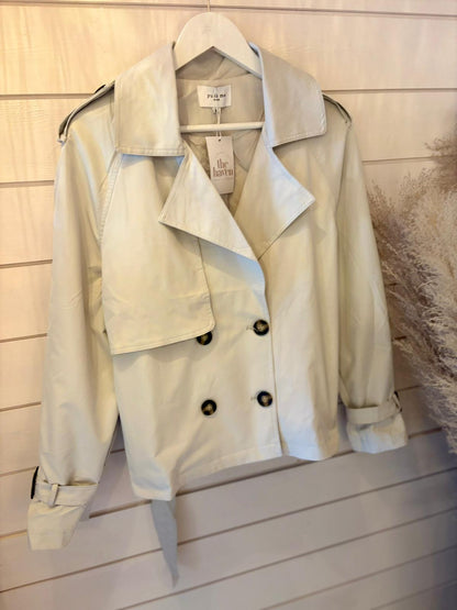 Light beige double-breasted trench coat with a wide collar and belted cuffs, shown on a hanger against a white wooden wall.
