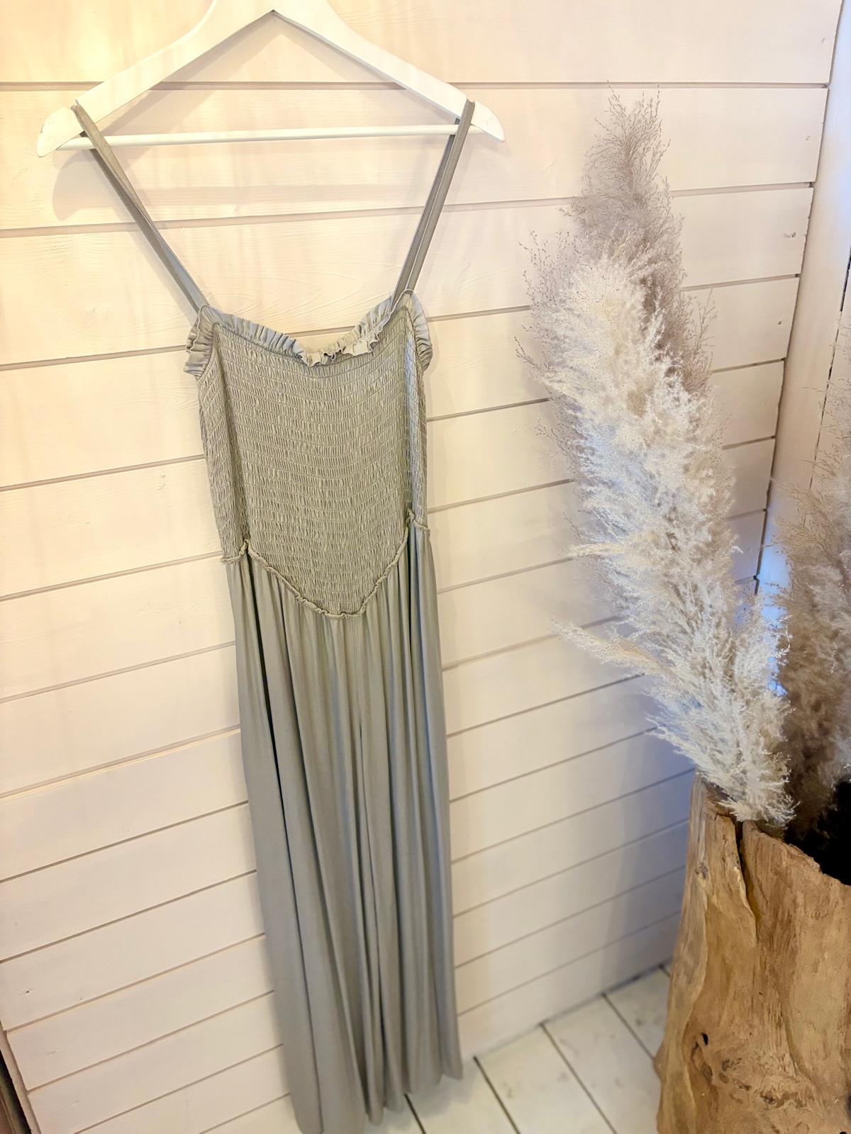 A light grey sleeveless maxi dress hangs on a white hanger against a wood-panelled wall, beside a large vase of pampas grass.
