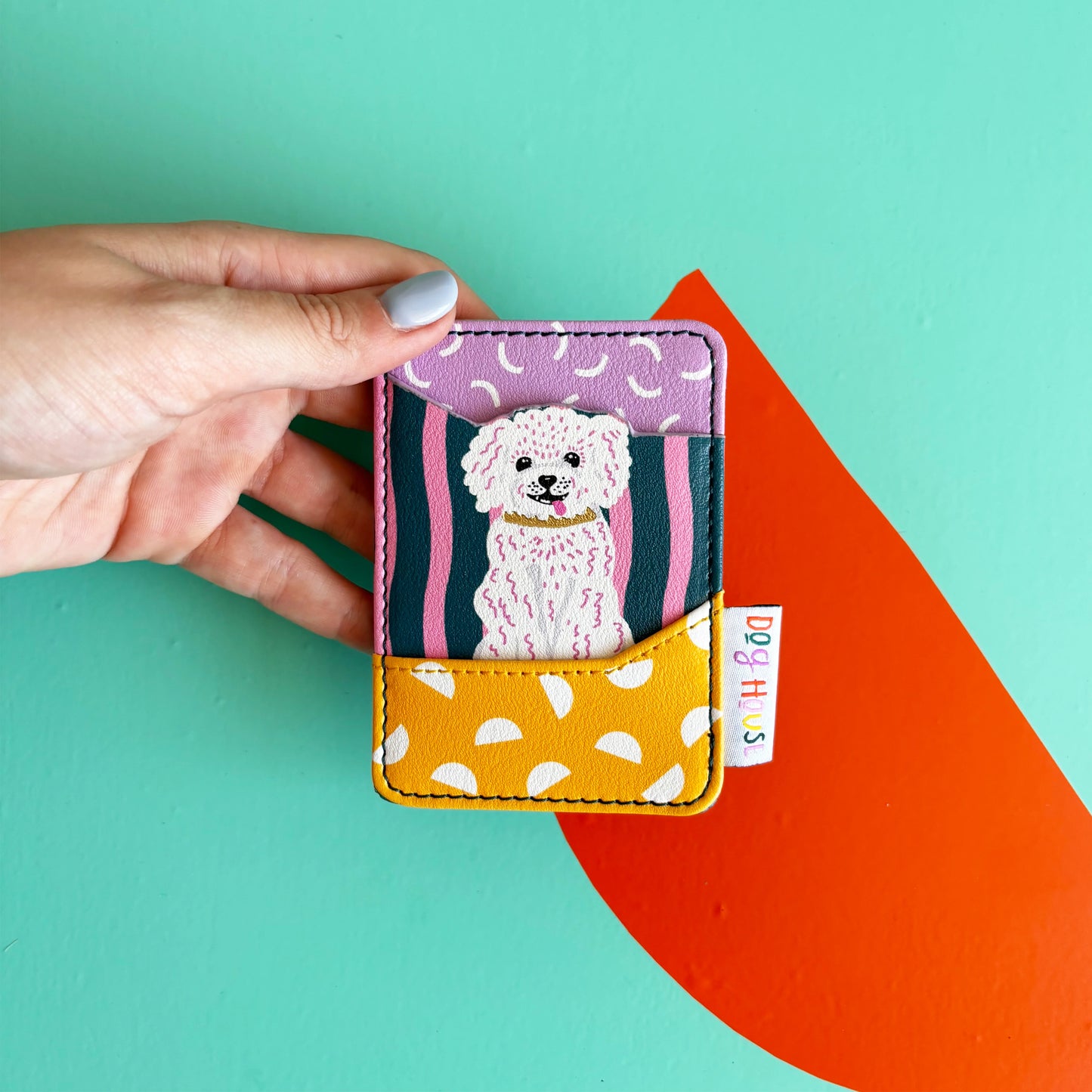 A hand holds a charming card wallet featuring a cartoon white dog illustration and colourful patterns, set against a teal background with an orange shape.