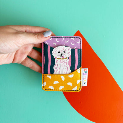 A hand holds a charming card wallet featuring a cartoon white dog illustration and colourful patterns, set against a teal background with an orange shape.