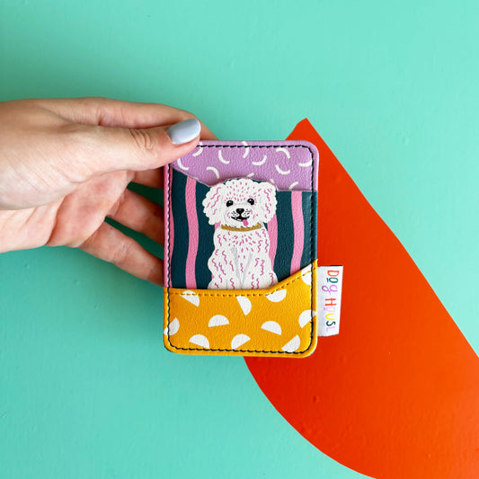 A hand holds a charming card wallet featuring a cartoon white dog illustration and colourful patterns, set against a teal background with an orange shape.