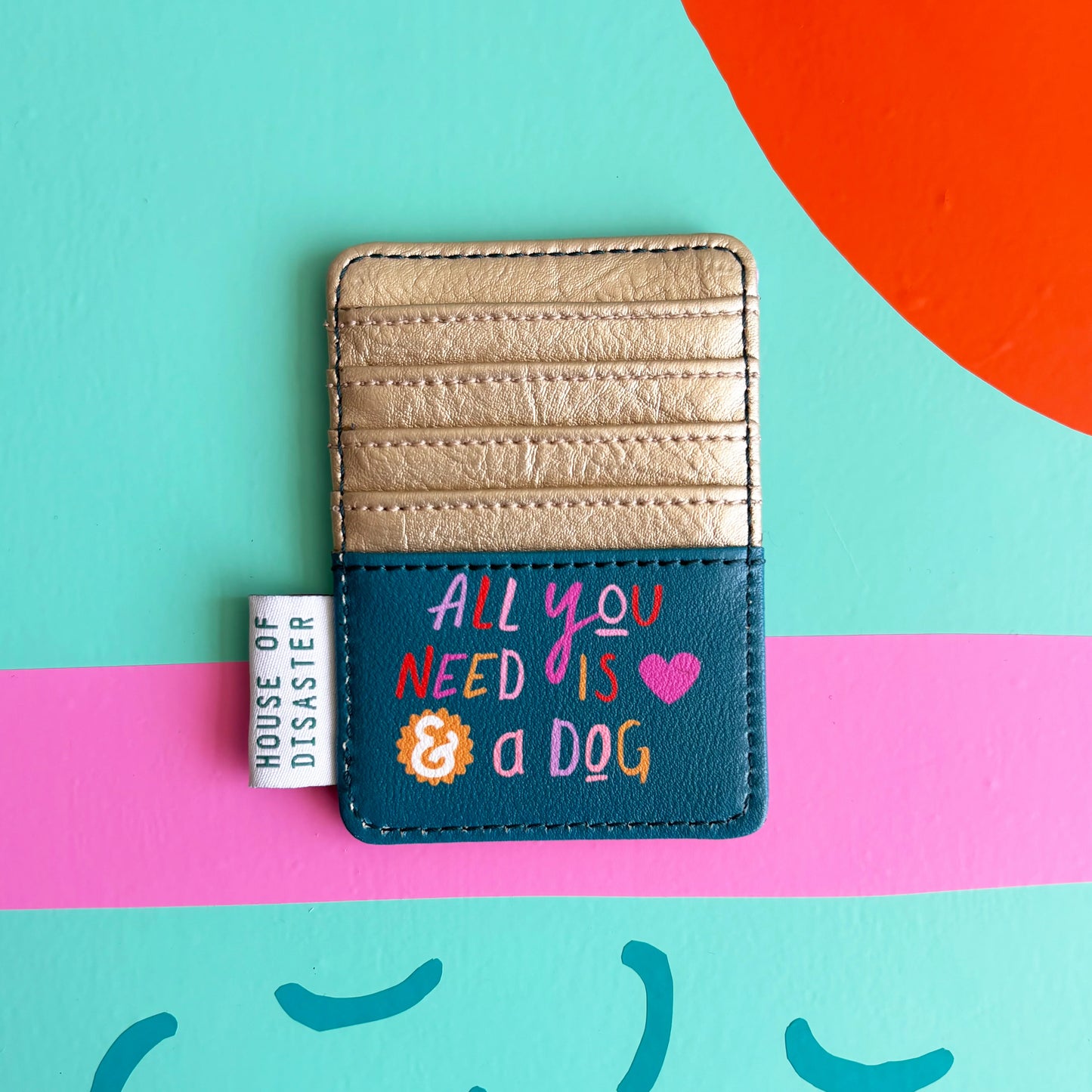 A charming card wallet with gold and blue panels reads “All you need is love & a dog” and features the House of Disaster tag, set against a colourful background.
