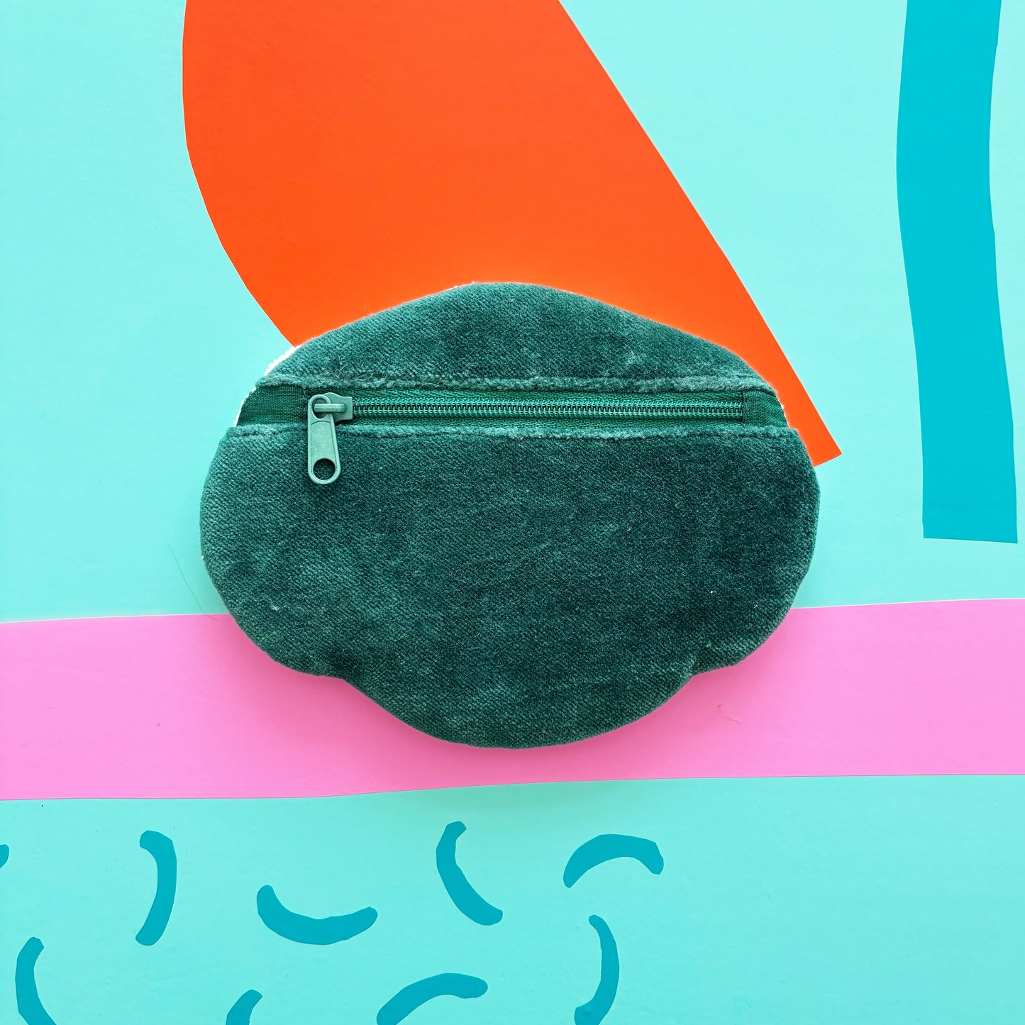 A round, green velvet coin purse with a zip rests on a colourful, abstract background featuring shapes in orange, pink, and blue.
