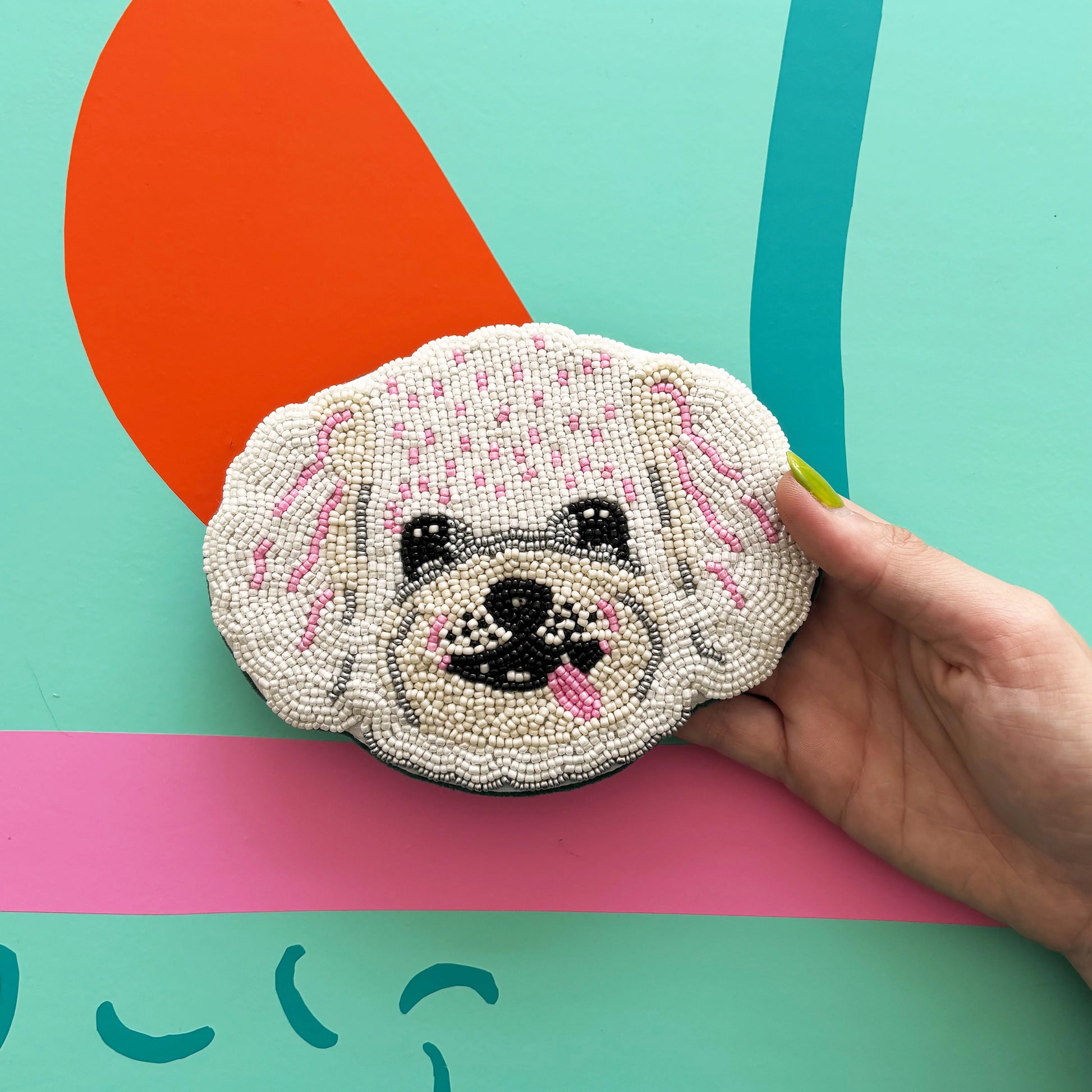 A hand holds a beaded coin purse shaped like a white dog's face with pink accents, set against a colourful abstract background.