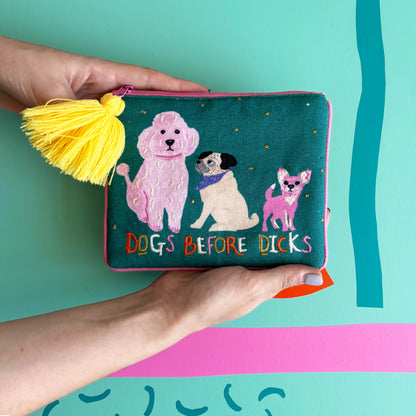 A person holds a quirky dog purse—a green velvet pouch with a yellow tassel, playful illustrations of three dogs, and the bold wording “DOGS BEFORE DICKS.”.