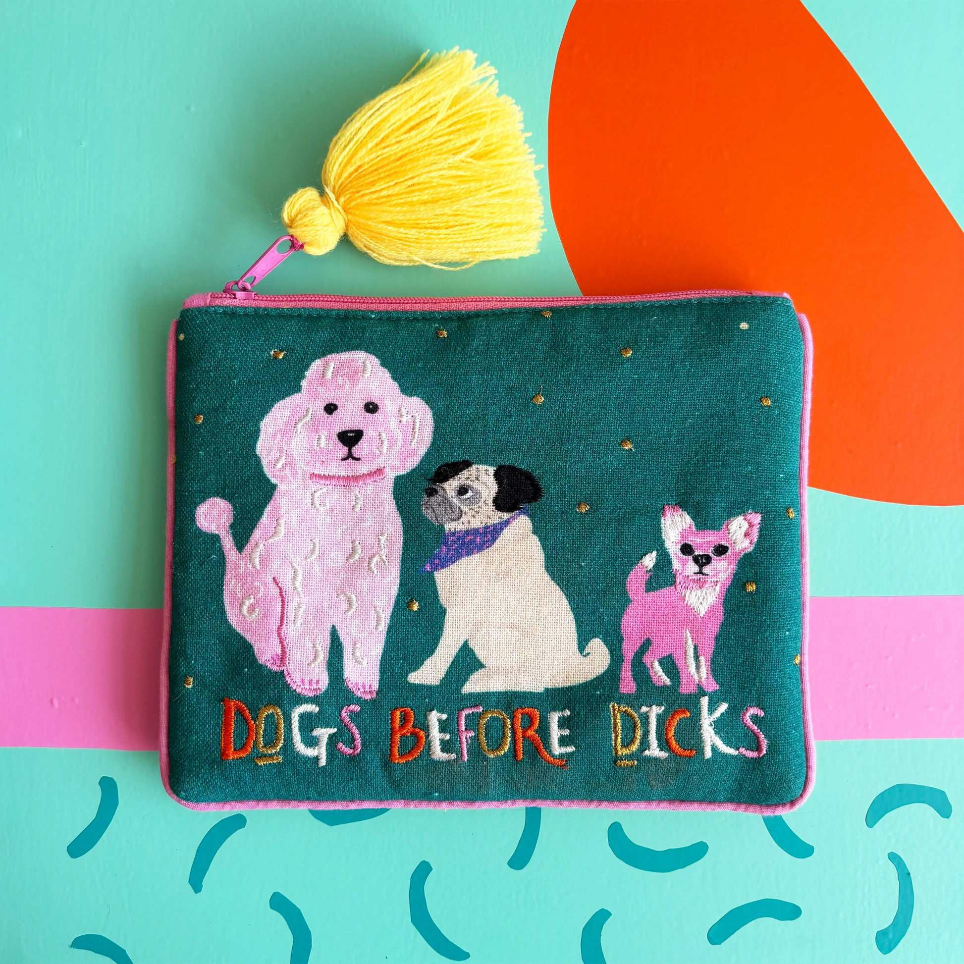A funky dog handbag, this green velvet pouch displays embroidered images of three dogs, the phrase DOGS BEFORE DICKS, and a yellow tassel, all set against a colourful abstract background.
