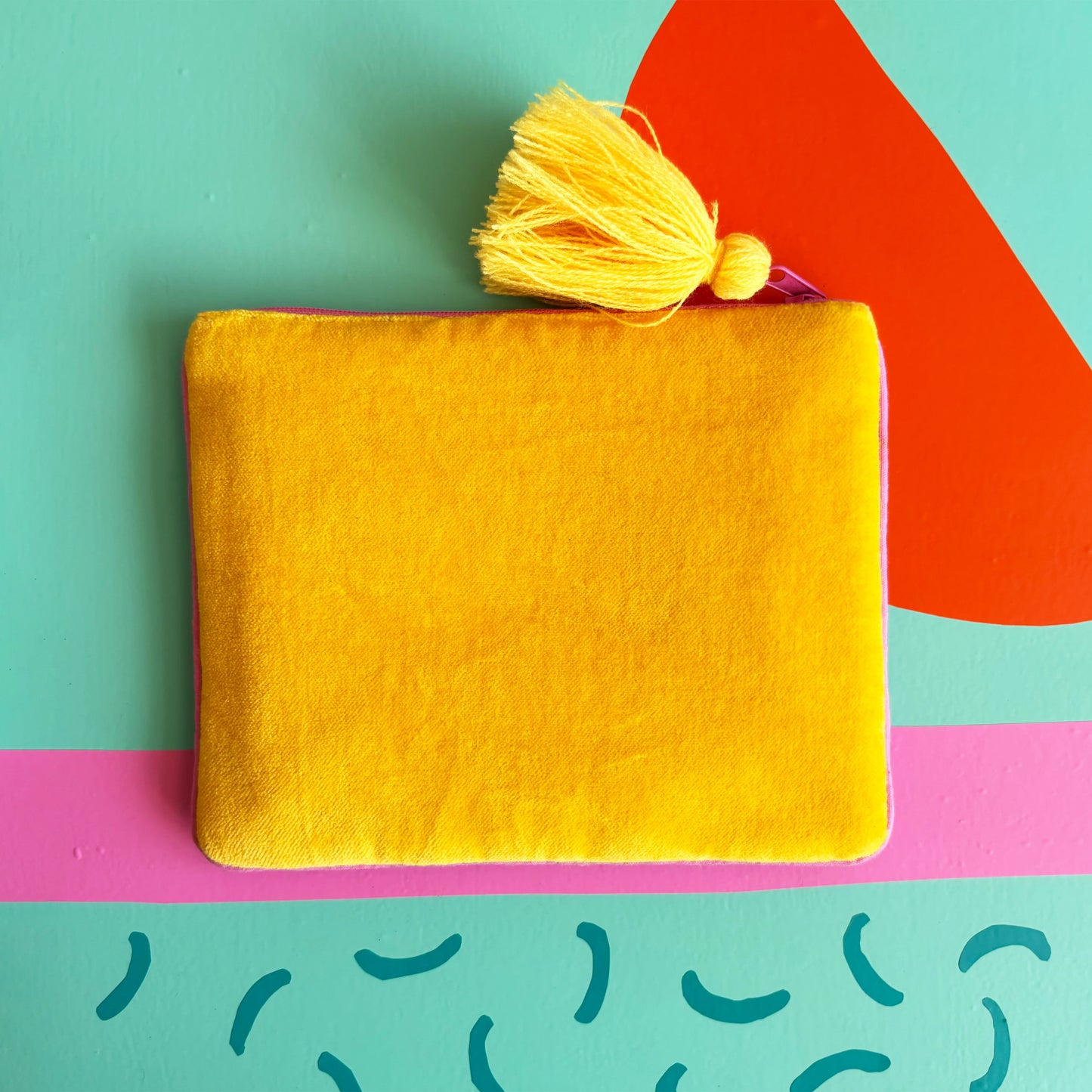 A yellow velvet handbag with a matching tassel rests on a colourful, abstract background featuring teal, pink, red, and blue shapes.