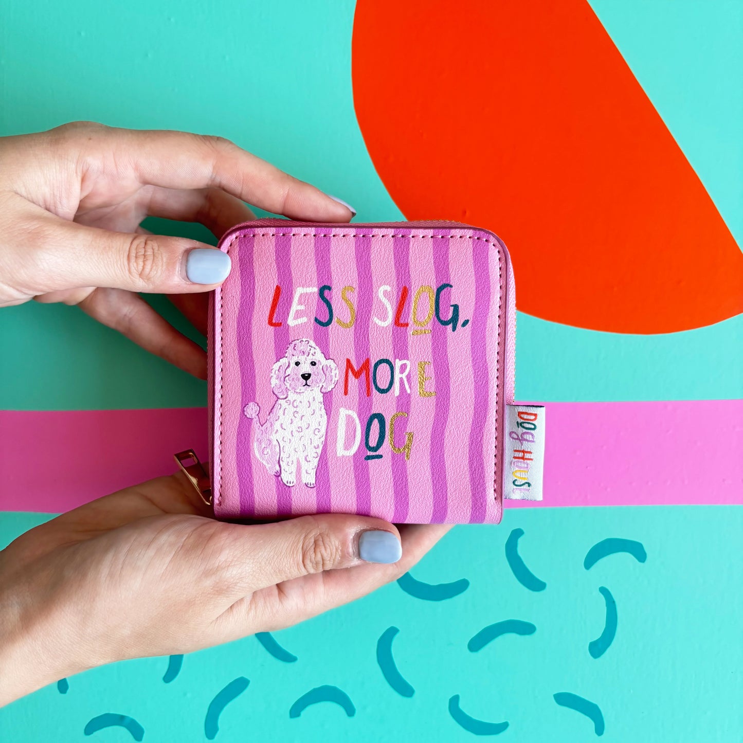 Two hands hold a pink striped zip purse from the Doghouse collection featuring a poodle illustration and the wording LESS SLOG, MORE DOG, set against a colourful abstract background—ideal for dog lovers.