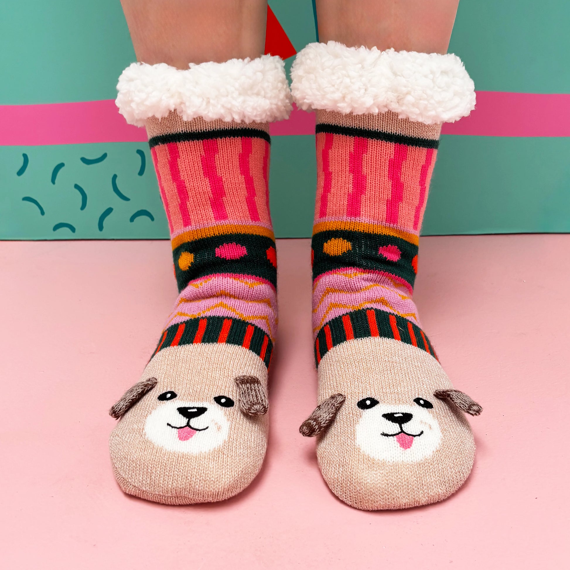 A person is wearing cosy, colourful Dog House Slipper Socks with a dog face design and fluffy cuffs, standing on a pink surface before a teal background—ideal as a gift for dog lovers.