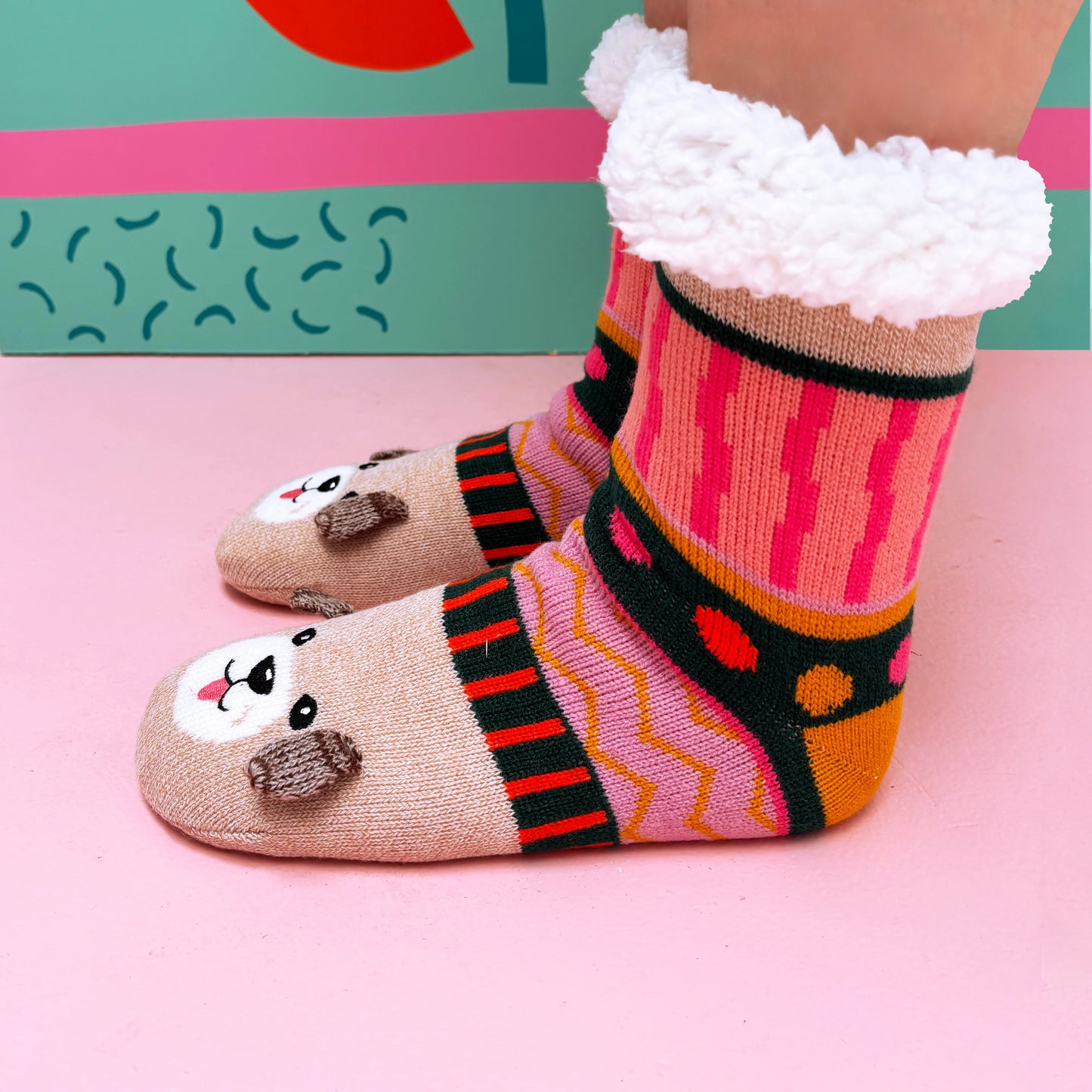 A person wearing colourful, patterned Dog House Slipper Socks featuring bear faces and fluffy white cuffs stands on a pink floor with a vibrant, colourful background.