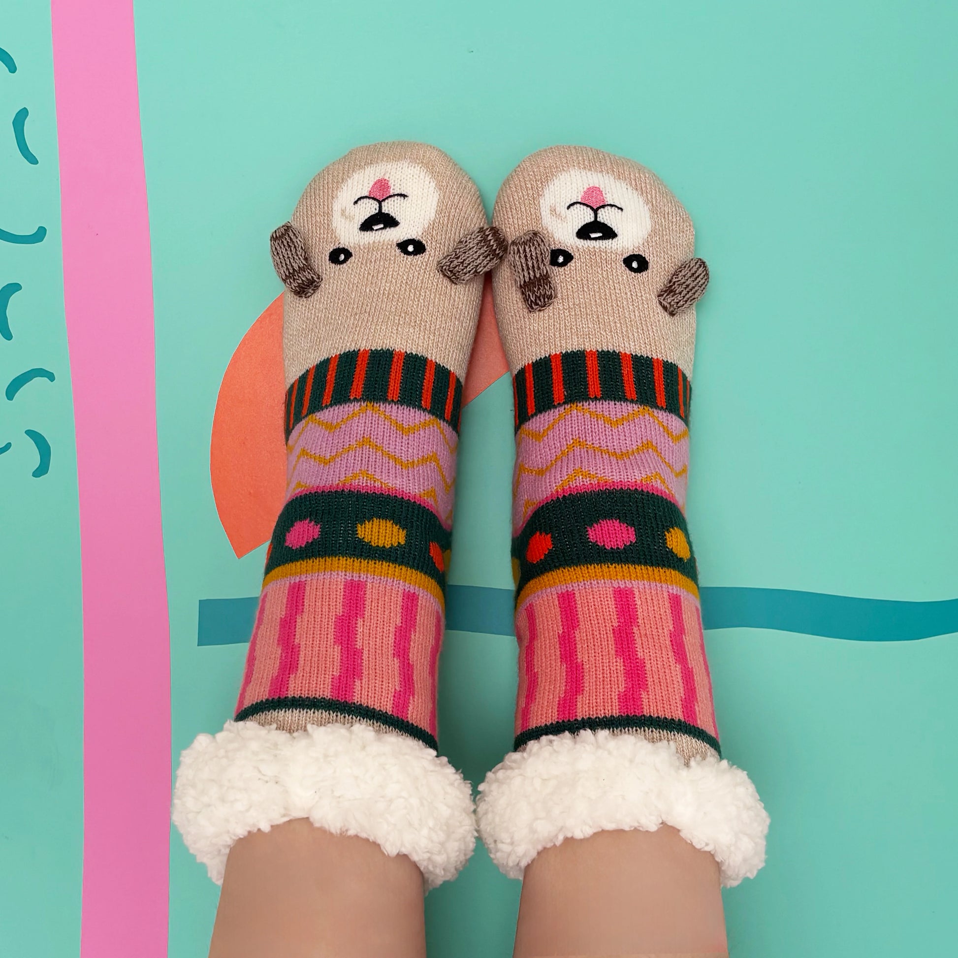 Two feet in colourful, patterned Dog House Slipper Socks with bear faces on the toes and fluffy white cuffs, featuring anti-slip soles—set against a teal background with abstract shapes.