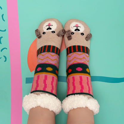 Two feet in colourful, patterned Dog House Slipper Socks with bear faces on the toes and fluffy white cuffs, featuring anti-slip soles—set against a teal background with abstract shapes.