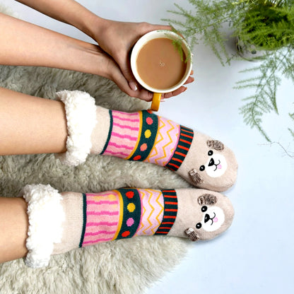 A person wearing colourful, patterned anti-slip slipper socks with bear faces holds a yellow mug of coffee or tea, seated on a light surface beside a green potted plant.